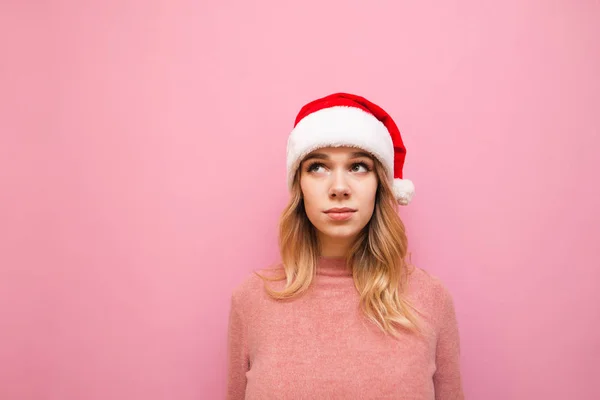 Portrait of a cute teenager girl in pink clothes and Christmas hat on a pink background, looking away at copy space.Blonde in santa hat stands isolated on pink background.Christmas and New Year concept — стоковое фото