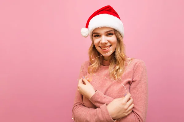 Szczęśliwa dziewczyna w kapeluszu Santa Claus i różowy sweter stoi na różowym tle ze światłami w ręku, patrzy w aparat i uśmiecha się. Odizolowany. Blondynka z girlandą w dłoni. Święta. Boże Narodzenie. — Zdjęcie stockowe