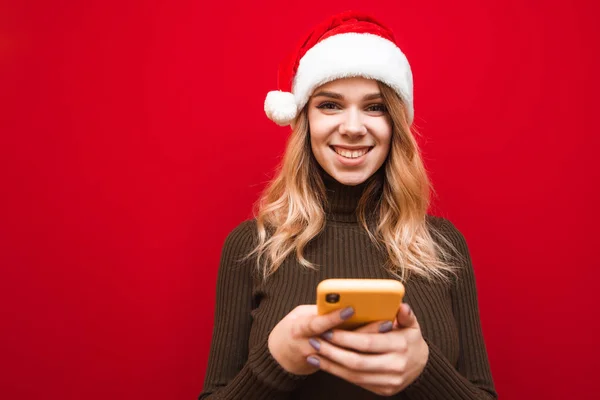 Ευτυχισμένη κυρία με καπέλο Χριστουγέννων απομονώνονται σε κόκκινο φόντο με smartphone στα χέρια κοιτάζοντας στην κάμερα και χαμογελώντας. Κοντινό πλάνο πορτρέτο του χαμογελαστού κοριτσιού των Χριστουγέννων με smartphone σε κόκκινο φόντο. — Φωτογραφία Αρχείου