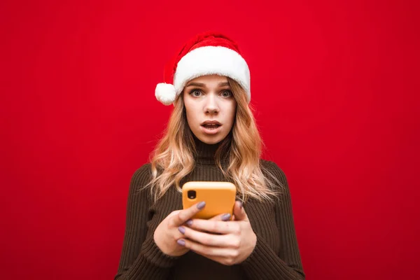 Förvånad flicka i jul hatt står på röd bakgrund med smartphone i händerna, chockad utseende ser in i kameran. Chockad dam i Santa claus hatt isolerad på röd bakgrund med smartphone i handen. — Stockfoto