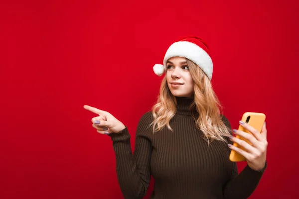 Menina atraente em chapéu de Papai Noel e suéter quente fica no fundo vermelho com smartphone na mão, olha e mostra polegares para cima no espaço de cópia. Conceito de Natal e Ano Novo . — Fotografia de Stock