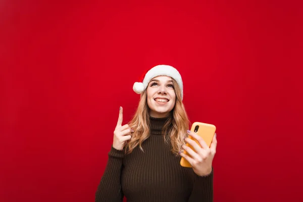 Радостная девушка в рождественской шляпе стоит на красном фоне со смартфоном в руке, смотрит и показывает пальцем вверх на пустое пространство. — стоковое фото