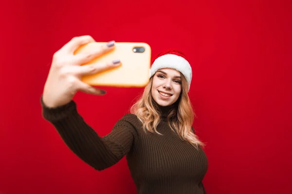 Привлекательная девушка в тёплом свитере на шляпе Санта Клауса делает селфи на смартфоне на красном фоне. Счастливая девушка в рождественской шляпе делает фото для себя. Рождественская селфи . — стоковое фото