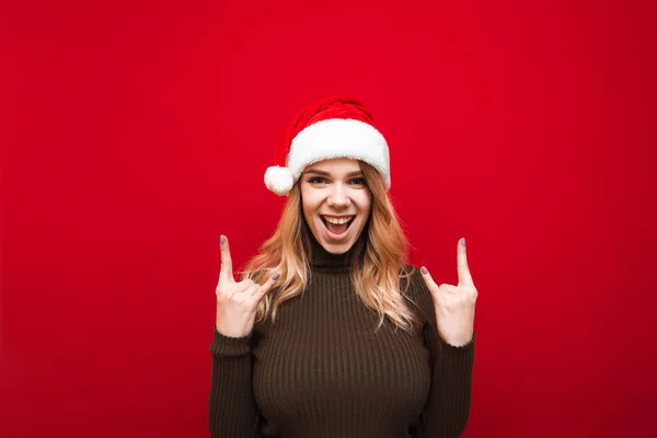 Glada flicka i Santa jul hatt visar heavy metal gest, tittar in i kameran och skrattar åt röd bakgrund. Flicka i tröja visar händer gest av året och ser känslomässigt med kameran — Stockfoto
