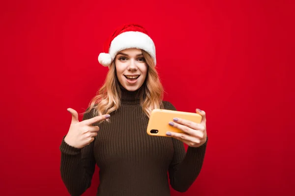 따뜻 한 스웨터를 입고 모자를 쓴 즐거운 소녀의 사진 이 빨간 배경에 서서 카메라를 들여다보며 미소를 짓고 손에 스마트폰을 들고 있다. 새롭고 크리스마스같은 개념. — 스톡 사진