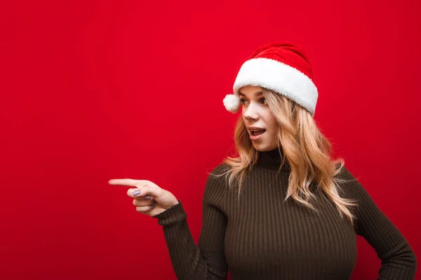 Menina chocada no chapéu de Natal isolado no fundo vermelho, surpreendido olhando para o espaço em branco e apontando o dedo para o espaço de cópia. Senhora atraente em Papai Noel, mostrando surpresa — Fotografia de Stock