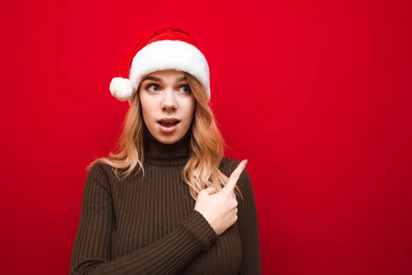 Retrato de senhora surpreso em chapéu de santa e suéter quente no fundo vermelho, mostra o dedo no espaço de cópia e olha para o lado com o rosto chocado. Isolado. Venda de X-mas — Fotografia de Stock