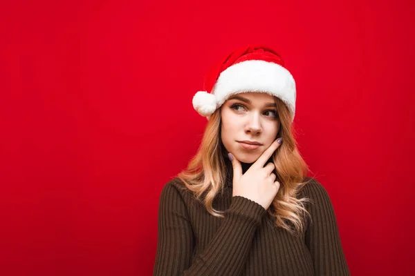 Smart tjej i tomte hatt och varm tröja isolerad på röd bakgrund, ser bort på kopieringsutrymme och tänker nära porträtt. Pensiv flicka i julhatt ser bort. Julkoncept. — Stockfoto