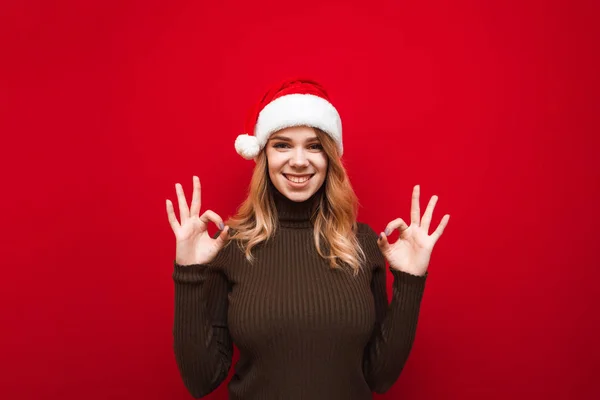 Happy girl in santa hat and warm sweater isolated on red background, hands showing OK gesture, looking into camera and smiling.Studio portrait of joyful lady in christmas hat showing gesture all right — стоковое фото