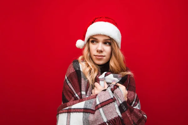 Menina loira bonita aquece em xadrez no fundo vermelho, usa chapéu de Papai Noel e olha para o lado. Retrato de uma menina congelada aquecendo em xadrez, isolado . — Fotografia de Stock