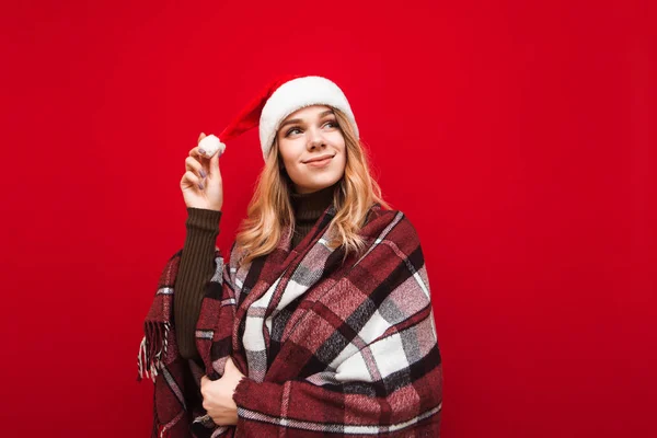 Positive girl in santa hat and warm plaid stands on red background and looks away with smile at christmas, isolated. Lady warms up in plaid on red background. Xmas concept — Stock Photo, Image