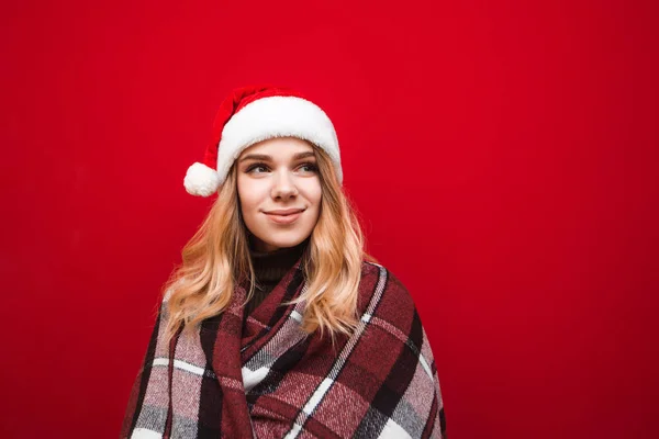 Привлекательная девушка в рождественской шляпе и клетчатой подогревается на красном фоне и отводит взгляд с улыбкой. Красивая дама в шляпе Санты и клетка в Рождество изолированы на красном фоне . — стоковое фото