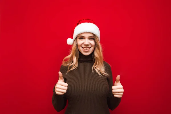 Menina feliz em suéter quente e chapéu Papai Noel fica no fundo vermelho, mostra os polegares para cima e olha para a câmera com sorriso, isolado. Dama de Natal positiva mostrando gesto como. X-mas — Fotografia de Stock