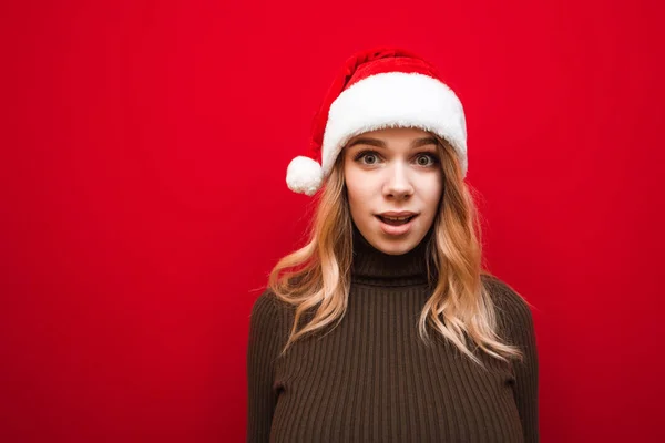 Närbild porträtt av överraskad vacker flicka i casual kläder, bär tomte hatt och varm vinter tröja, chockad tittar in i kameran på röd bakgrund. Chockad dam i jul hatt isolerad. — Stockfoto