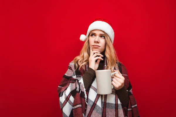 Ragazza bella riflessivo in un plaid caldo con una tazza di bevanda calda in mano si erge su uno sfondo rosso, guarda di lato e su e pensa. Ritratto natalizio di una ragazza con una tazza di tè . — Foto Stock