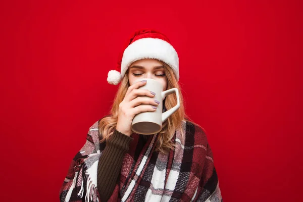 Ragazza carina a Babbo Natale cappello e bevande plaid bevanda calda da grande tazza con gli occhi chiusi e gode su sfondo rosso. Lady beve il tè e si riscalda in plaid in inverno. Concetto Natale . — Foto Stock