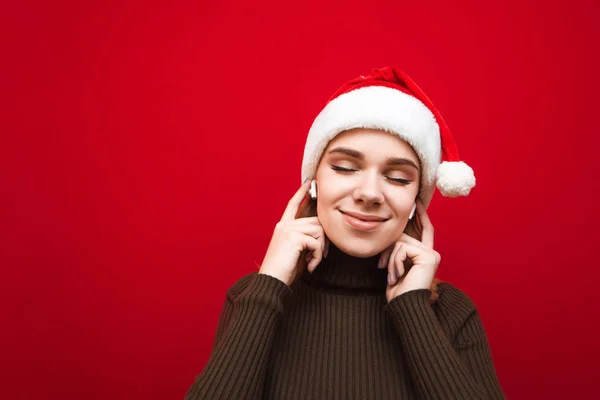 Närbild porträtt av attraktiv flicka njuter av musik med slutna ögon i trådlösa hörlurar över röd bakgrund. Positiv tjej i tomteluva som lyssnar på musik i hörlurar. Isolerad. — Stockfoto