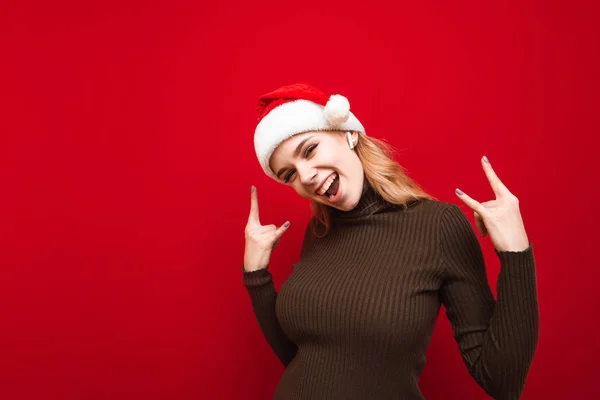 Радостная девушка в шляпе Санты слушает рок-музыку в беспроводных наушниках на красном фоне, смотрит в камеру, улыбается и показывает руки с жестом хэви-метала. Xmas — стоковое фото