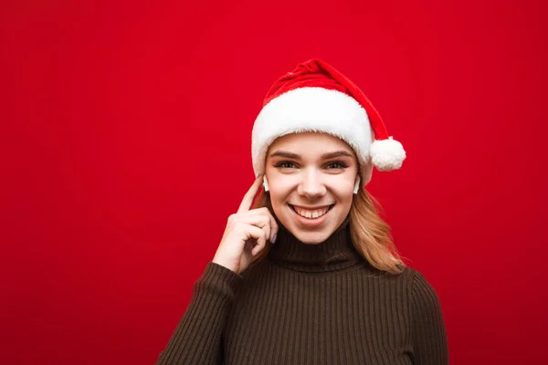 Крупный план фотографии улыбающейся девушки в шляпе Санты, стоящей на красном фоне в беспроводных наушниках, смотрящей в камеру и улыбающейся на красном фоне. Леди слушает рождественскую музыку, изолированную — стоковое фото