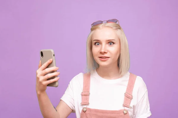 Chica emocional con un teléfono inteligente en sus manos sobre un fondo púrpura mira a la cámara con asombro. Retrato de cerca de una adolescente sorprendida con un teléfono inteligente en la mano. Aislado . — Foto de Stock