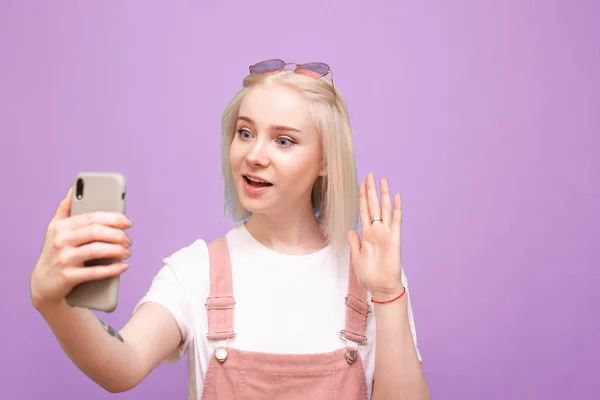 Dulce chica con un teléfono hablando en una videollamada en el teléfono inteligente, mirando a la pantalla del teléfono con la cara asombrada sobre un fondo púrpura. Adolescente chica hace selfie en un rosa fondo . — Foto de Stock