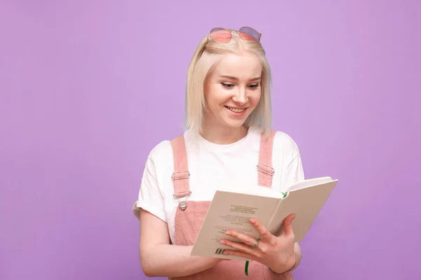 Ευτυχισμένο ξανθό κορίτσι με ένα βιβλίο στα χέρια της σε μωβ φόντο χαμογελάει και διαβάζει. Ευτυχισμένο έφηβο κορίτσι με χαριτωμένα ρούχα που διαβάζει ένα βιβλίο και γελάει, απομονωμένο σε ροζ. — Φωτογραφία Αρχείου