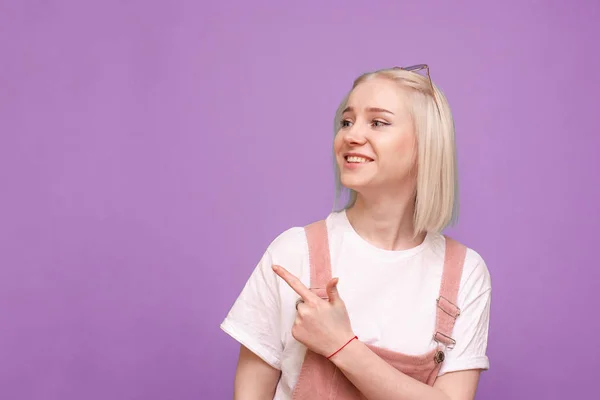 Ragazza felice in abiti carini in piedi sullo sfondo di un muro viola, mostrando il dito da parte e sorridente. Teen girl distoglie lo sguardo e mostra il dito sul copyspace. Isolato su sfondo rosa . — Foto Stock