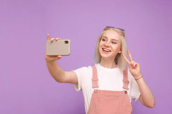 Menina bonito feliz em uma roupa casual leve faz selfie no fundo de uma parede roxa, olha para o smartphone e sorri. A adolescente faz uma foto ao telefone. Isolados . — Fotografia de Stock