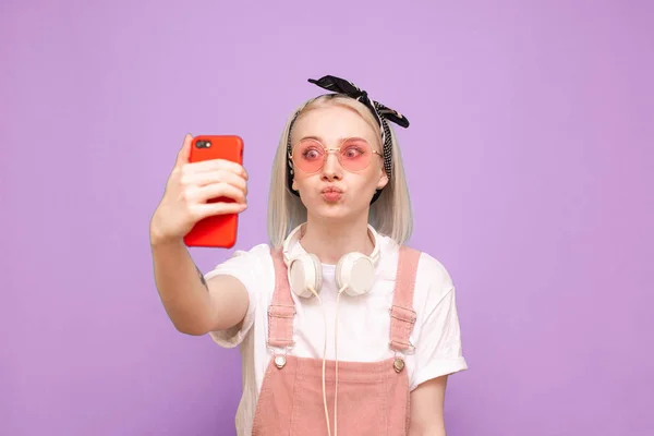 Chica divertida en ropa linda toma una selfie con una cara divertida sobre un fondo púrpura. Divertida adolescente chica hace selfie en un teléfono inteligente, con ropa brillante y gafas de sol de color rosa — Foto de Stock