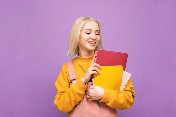 Étudiante heureuse aux cheveux clairs et aux vêtements décontractés se tient debout sur un fond violet, tient un cahier à la main et sourit. Portrait d'un étudiant souriant avec des livres, isolé, concept d'apprentissage . — Photo