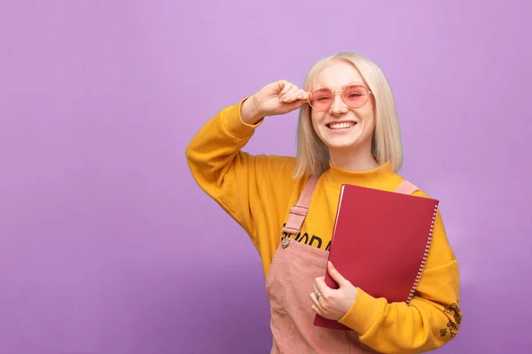 Portret van een vrolijk studentmeisje met een roze bril en een notitieblok in haar hand staat op een paarse achtergrond, kijkt naar de camera en glimlacht. Stijlvol meisje met boeken in haar handen glimlachend, geïsoleerd. — Stockfoto