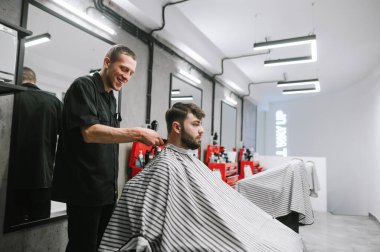 A smiling barber cuts a customer with a clipper in a bright modern barber shop. Happy male hairdresser creates hairstyle to bearded man. Positive barber uses clipper.