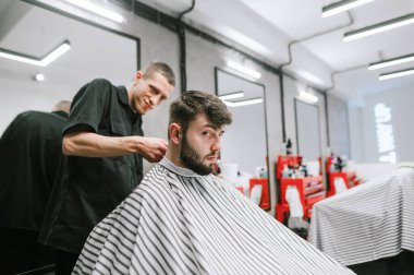 Portrait of a man sitting in a chair in a men's hairdresser and looking into the camera, smiling hairdresser doing hairstyle to client. Positive barber cuts a man with a serious suspicious face