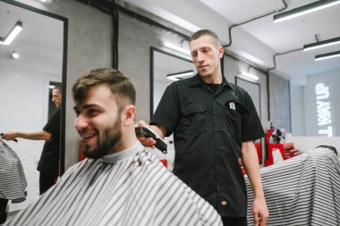 A male hairdresser cuts a smiling client with a bororo, a hairdresser uses a clipper. Portrait of a barber cuts a man with a clipper in his hand, looks at his hair. Creating a haircut in a barbershop