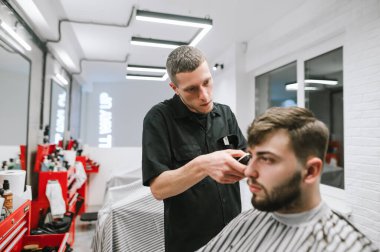 Concentrated barber makes hair bearded client in a modern light barber shop, uses a clipper. Male hairdresser prepares hairstyle for man in hairdresser.