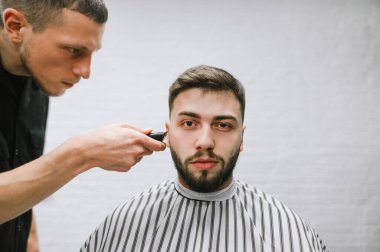 Man with a beard visits a barber shop, a barber repeats creating a hairstyle with a clipper in his hands. Professional hairdresser makes stylish hairstyle for a beautiful client in a male hairdresser