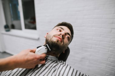 Barber trims the beard of a handsome man with a clipper in hand. Closeup portrait of barber shop client. Barber correction of client's beard. Hairdresser doing hairstyle to bearded client in barbersho