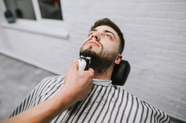 Professional barber with a hair clipper in his hand makes a beard hairstyle for the client. Man receives a service in a barber shop, hairdresser is engaged in the correction of his beard.