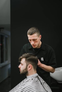 Concentrated barber cuts the client's scissors in a dark barber shop. Male hairdresser creates a stylish hairstyle for a bearded man, sitting in a chair with a serious face. Vertical