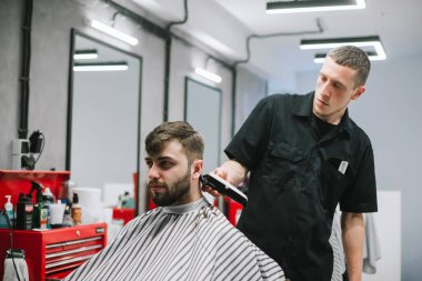 Barber trims client's hair with a clipper in his hands. Barber works in a barbershop workplace, makes a stylish hairstyle for a bearded man. Barber job concept.