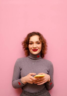 Portrait attractive positive girl with makeup and curly red hair on pink background, uses smartphone and looks into camera with smile on face,isolated. Vertical portrait of happy girl with smartphone
