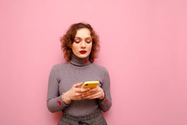 Beautiful girl with curly hair wears a gray sweater, stands on a pink background and uses a smartphone. Cute teen girl writes a message on smartphone, isolated. Copy space