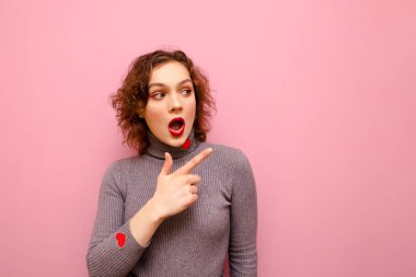 Surprised young girl in casual clothes and red curly hair shows pointing finger at copy space. Cute emotional lady showing thumbs up with surprised face on pink background.
