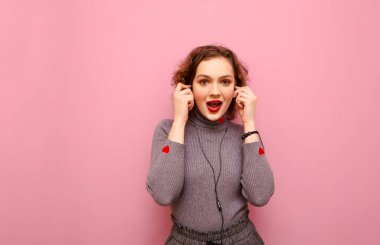 Cheerful girl with red curly hair listens to music in headphones on a pink background, looks into the camera with a surprised face. Lady with bright makeup and gray sweater listens to music, isolated