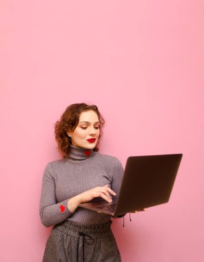 Vertical portrait of a curly red-haired girl in gray casual clothes uses a laptop on a pink background, looks into the screen with a serious face. Lady freelancer working on laptop ,isolated.