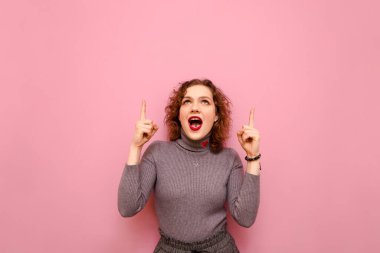 Emotional cute teen girl with curly red hair stands on a pink background and looks and shows thumbs up on copy space. Surprised lady in casual clothes shows up on empty space, isolated.