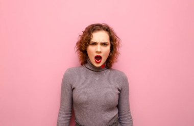 Portrait of emotional cute curly girl with red hair isolated on pink pastel background, looking into camera with surprised face. Shocked stylish lady in gray sweater stands on pink background.