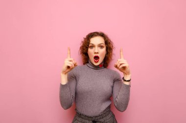 Portrait of shocked cute girl in casual clothes and with curly hair on pink background, shows thumbs up in blank space and with surprised face looks into camera. Cppy space