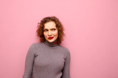 Cute girl with a suspicious look and curly red hair isolated on a pink pastel background, looks away at copy space with a thoughtful face. Isolated.