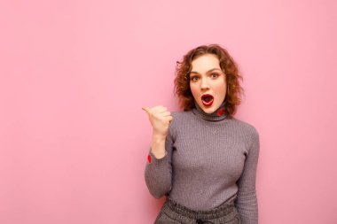 Cute surprised girl in a gray sweater and curly hair points her finger to the side and looks into the camera with a shocked face.Beautiful lady with red hair shows thumb up at copy space and is amaze